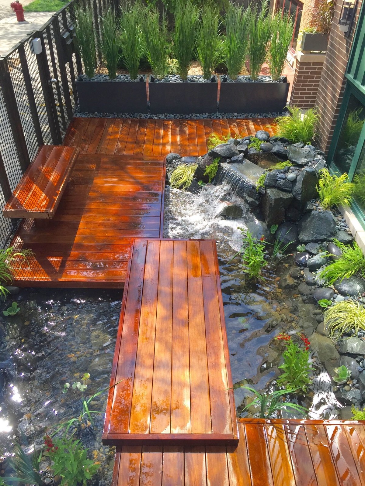 Wooden deck and boardwalk over a stream with large planters and rocks in a backyard patio